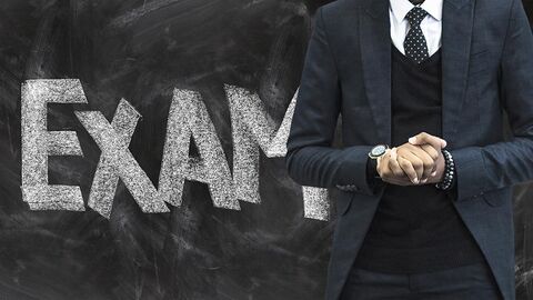 Mann (nur der Rumpf zu sehen) steht vor einer Tafel, auf der die Buchstaben EXAM zu erkennen sind.
