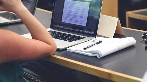 Tisch mit Laptop, Block und Stift; davor sitzt eine Person, von der nur ein Arm sichtbar ist.